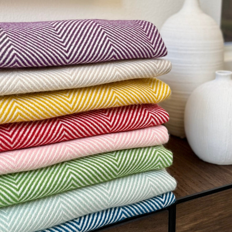 Stack of colorful herringbone-patterned fabrics on a wooden surface with white vases in the background.