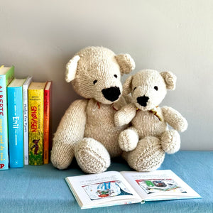 Two knitted teddy bears and an open book on a blue surface with books in the background.